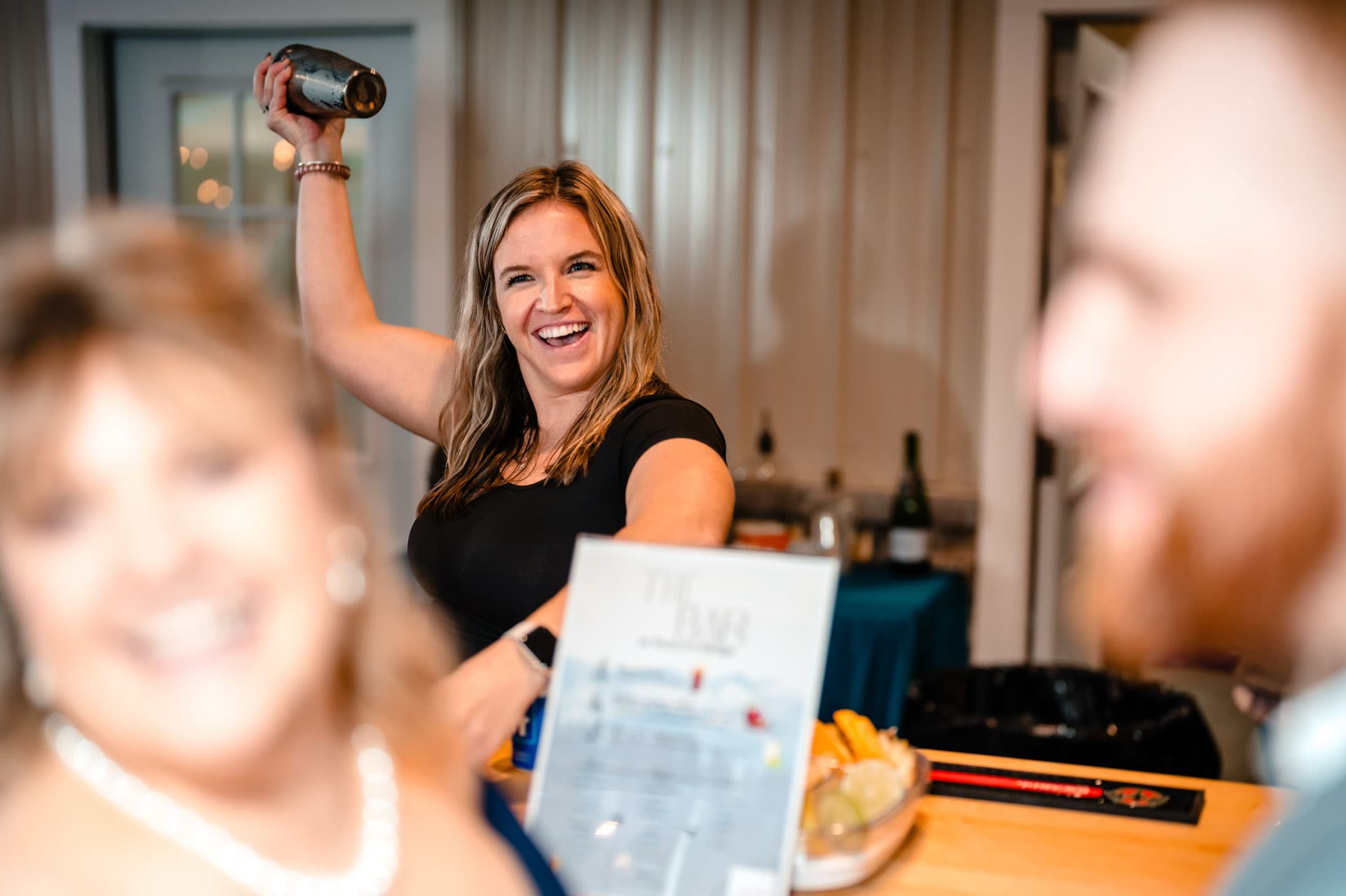Kayla raising a cocktail shaker behind the bar as wedding guests smile