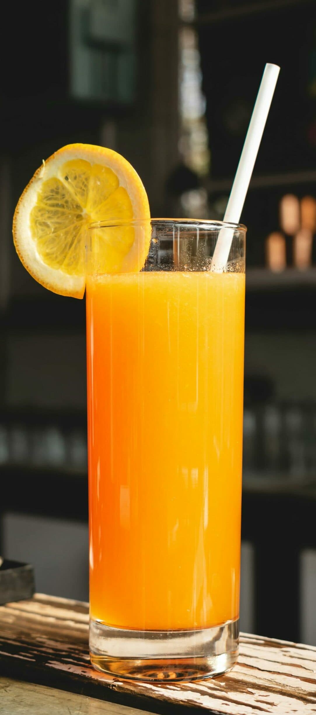 A tall orange drink with a lemon wheel and straw on a wooden bar