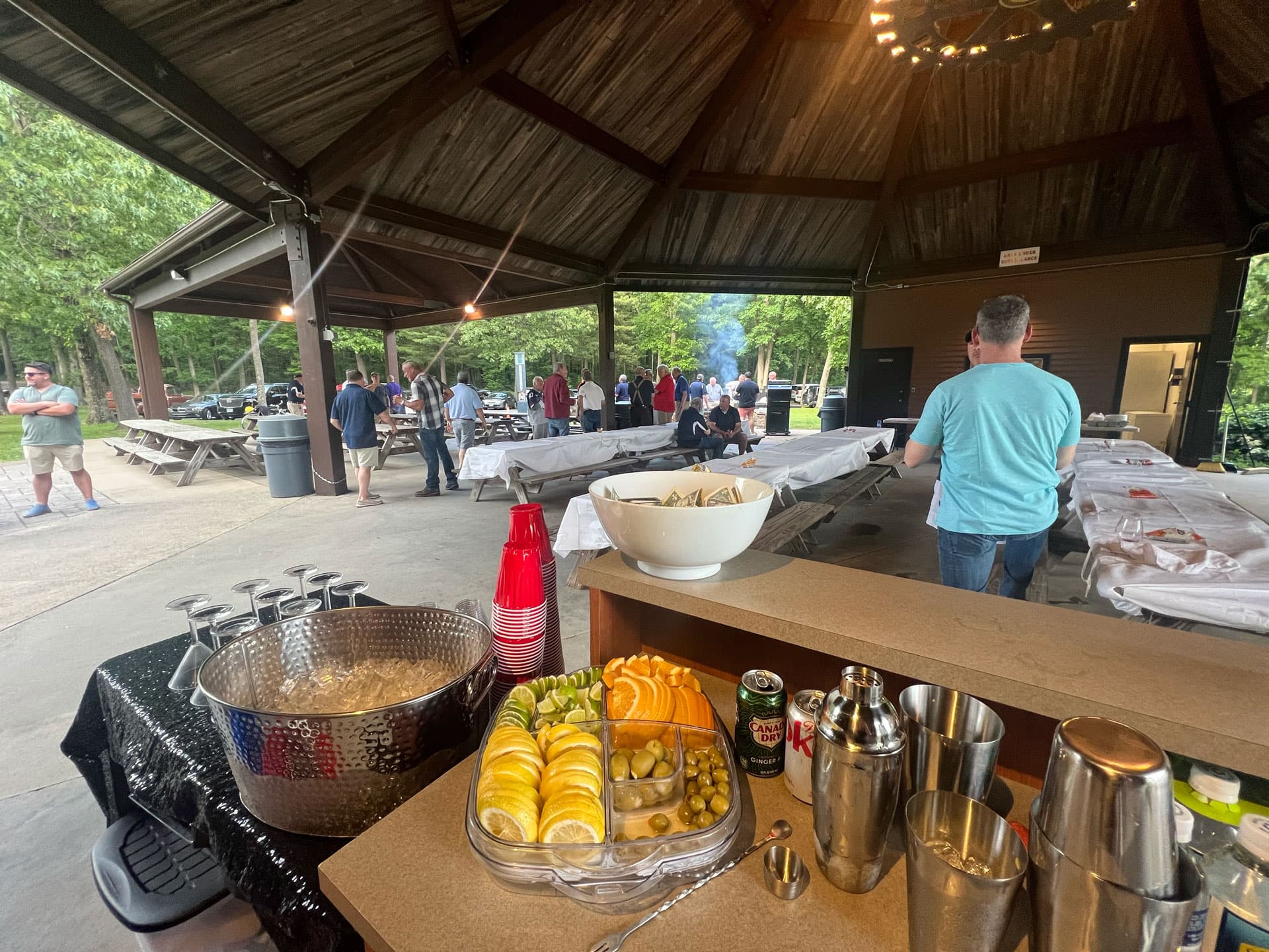 A bar set up under an outdoor pavilion with fruit, shakers, and guests