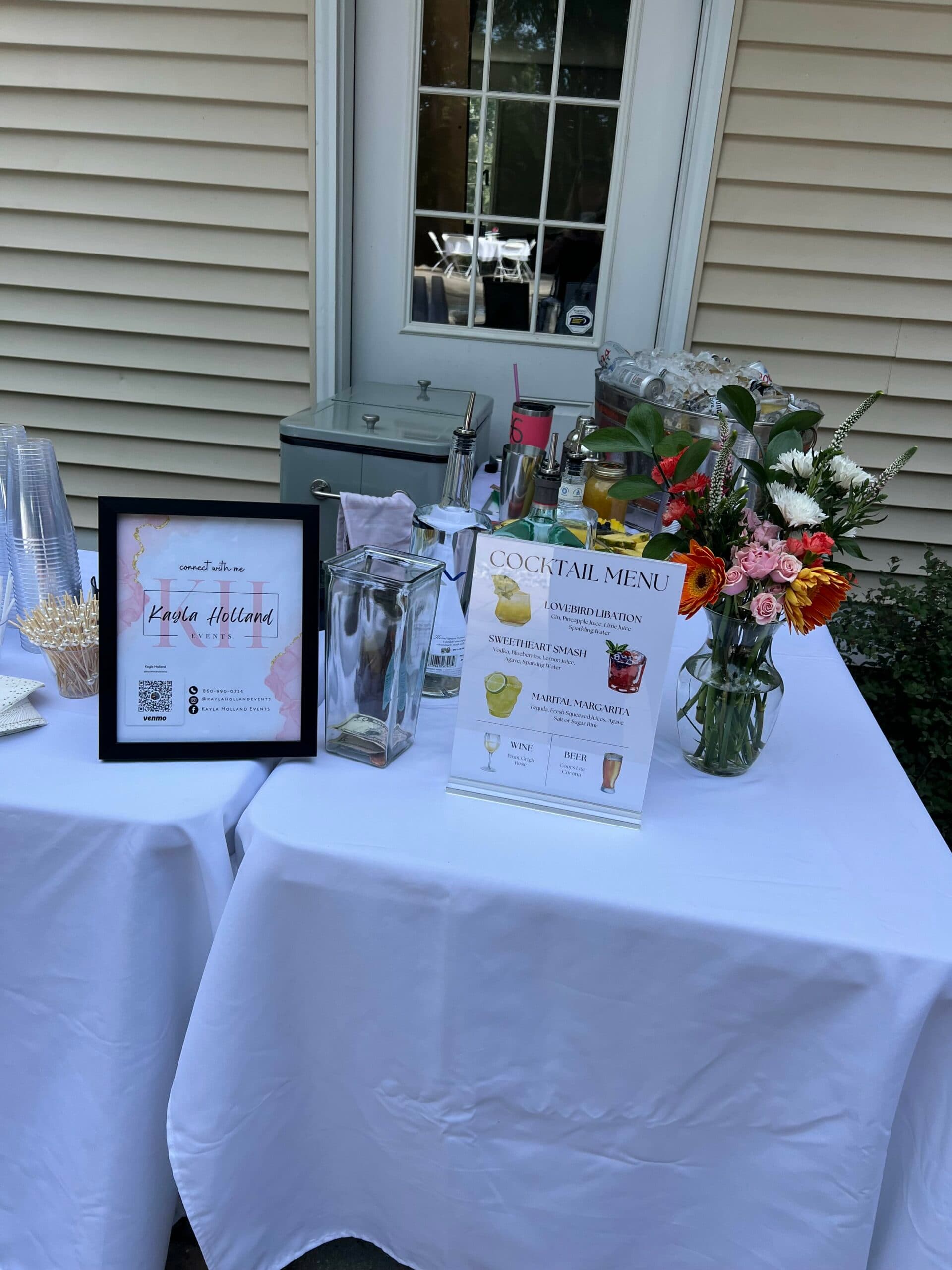 A welcome table with a framed sign, cocktail menu, and flowers on a white cloth